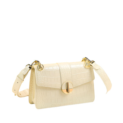 A milky white, crocodile patterned women's crossbody bag with wide straps and a zipper closure, displayed in front of a decorative background.